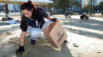 mulher está agachada na faixa de areia recolhendo microlixo e depositando em saco de papel. #paratodosverem