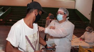 #pracegover Em ginásio, enfermeiro segura algodão no braço de homem vacinado
