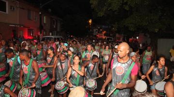 Integrantes de banda, com instrumentos de percussão e uniformizados estão na rua. #Pracegover