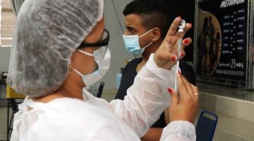 Enfermeira prepara vacina e jovem ao fundo. #paratodosverem