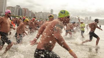 homens entram no mar para prova aquática. Eles estão correndo na beira da água indo em direção ao fundo para nadar. #paratodosverem 