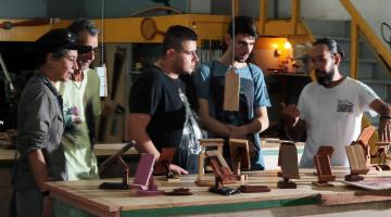 cinco pessoas estão em pé diante de uma mesa com peças em madeira. um deles, o professor fala ao grupo