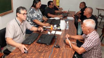 pessoas estão sentadas em torno de mesas. De um lado, o pessoal de atendimento, com computadores abertos. Do outro, pessoas fazendo consultas. #paratodosverem