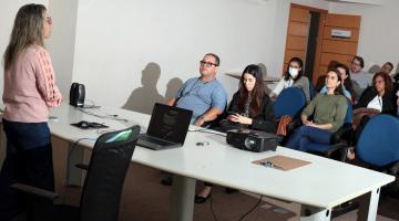 pessoas sentadas em auditório assistindo palestra. Uma mulher está em pé atrás de uma mesa falando a todos. #paratodosverem