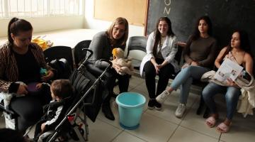 mulher segura boneco próximo a um balde. Mães em torno orbservam. #paratodosverem