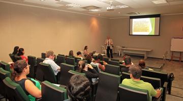 Auditório com servidores sentados assistindo palestra. Ao fundo aparece na parede uma imagem projetada. #Pracegover