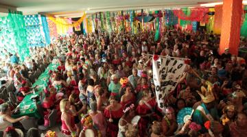 Salão de festas decorado para carnaval lotado de pessoas fantasiadas e dançando. #Pracegover