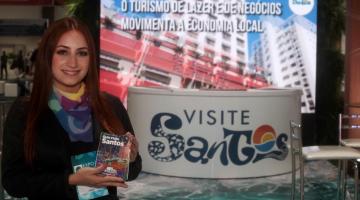 mulher segura panfleto em estande de santos de turismo #paratodosverem