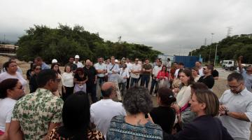 pessoas em pé em grupo num canteiro de obras, com morros ao fundo. #paratodosverem 