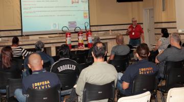 pessoas sentadas em aditório assistem palestra de um homem em pé. Ao fundo há um telão com informações e, pouco à frente, mesa com três extintores. #paratodosverem 