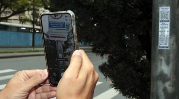 mãos seguram celular em cjua tela se fotografa o adesivo e aparece a placa da zona azul afixada em um poste que aparece à direita. #paratodosverem 