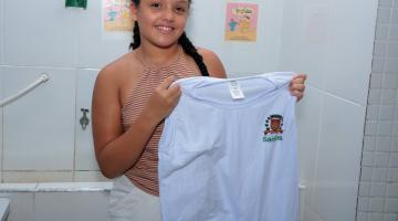 menina sorri para foto segurando camiseta de uniforme aberta pelas duas mãos. #paratodosverem 