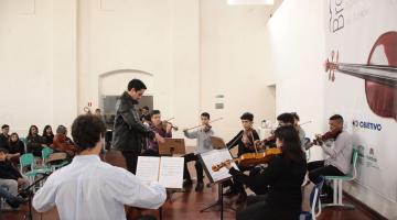 Jovens estão em uma sala, sentados e tocando instrumentos. Alguns têm as estantes à sua frente, com as respectivas partituras. #Pracegover