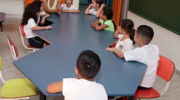 crianças pequenas, en torno de cinco anos, estão sentadas em torno de msa atentas à professora, que está na cabeceira, #paratodosverem 