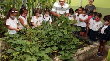 jardineiro mexe na horta e crianças observam #paratodosverem