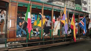 bonde com pessoas carregando bandeiras de vários países por fora do carro. #paratodosverem 