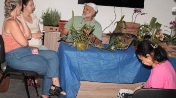 homem etá sentado atrás de uma mesa onde há várias mudas de plantas. Duas mulheres sentadas ao lado conversam com ele. #paratodosverem 