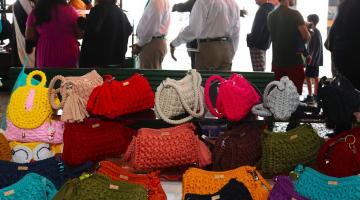 bolsas de crochê sobre mesa de exposição con pessoas circulando ao fundo. #paratodosverem 