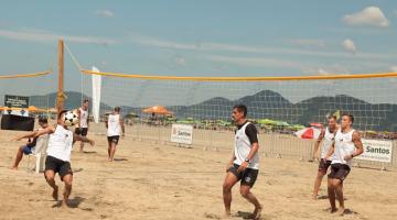 jovens estão jogando futevôlei na areia. Um deles aparece com a bola à frente do rosto, próximo ao peito, em movimento. Outros estão atentos à bola. #paratodosverem 