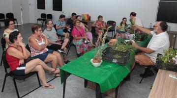 pessoas estão sentadas em auditório assistindo a um momente que fala sentado diante de uma mesa sobre a qual há vários vasos de orquídeas. #paratodosverem 