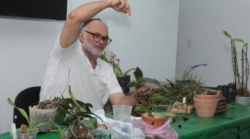 homem está sentado atrás de uma mesa com várias mudas de orquídeas e vasos. #paratodosverem 