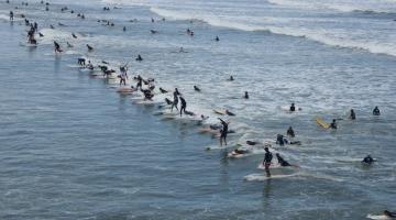 surfistas estão sobre uma mesma onda no mar em Santos. #paratodosverem 