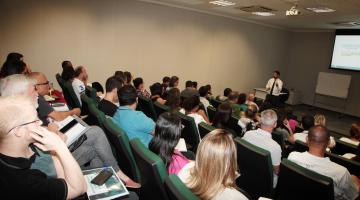 sala ampla com várias pessoas sentadas , à frente uma tela #paratodosverem