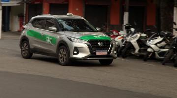 táxi passando por uma rua. Atrás há motos estacionadas junto ao meio-fio. #paratodosverem 
