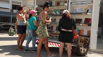 estande com livros expostos em praça. Pessoas manuseiam exemplares. #paratodosverem 