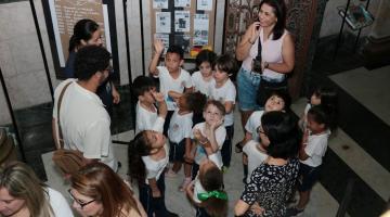 crianças reunidas no saguão do paço municipal diante de paineis com imagens de trabalhos escolhares. #paratodosverem 