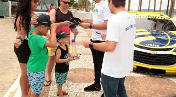 veículo da stock car ao fundo e pilotos conversando com o público à frente. #paratodosverem