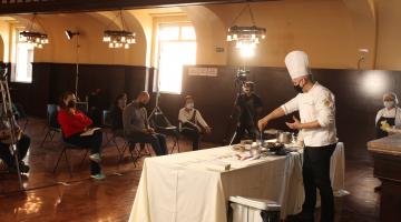 #pracegover Com uniforme de chef de cozinha, Júnior Monteiro maneja talheres sobre mesa à frente de câmeras 