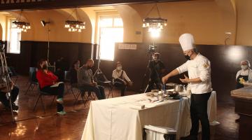 #pracegover Com uniforme de chef de cozinha, Júnior Monteiro maneja talheres sobre mesa à frente de câmeras 