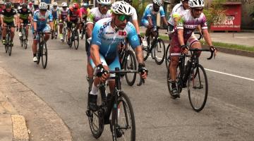 ciclistas estão pedalando em avenida. eles usam roupas de competição e capacete. #paratodosverem