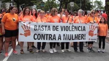 mulheres vestidas com camiseta na cor laranja estão em avenida e as da frente seguram uma faixa onde se lê basta de violência contra a mulher!#paratodosverem