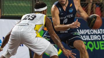 atleta de santos está de frente para a imagem. Ele está com a bola na mão esquerda, preparando para bater no chão. À frente dele, o adversário está com as pernas abertas, em pé e como tronco inclinado para baixo, em posição de contra-ataque. #paratodosverem