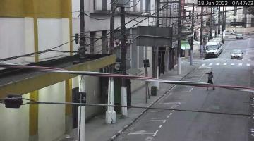 imagem a partir de câmera de monitoramento. No meio da rua se vê um homem caminhando e segurando peças nos ombros. #paratodosverem