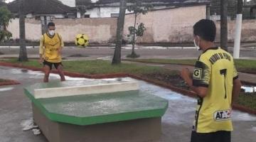 futmesa no centro de uma praça com dois jovens jogando bola, um em cada lado da estrutura. #paratodosverem