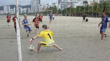 trave de gol em primeiro plano. Atrás,o goleiro tenta defender uma bola. Jogadores estão diante dele olhando a bola entrar na rede. Jogo é na praia. #paratodosverem