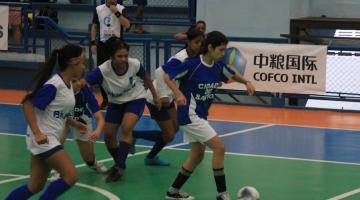 Meninas de uniformes branco e azul jogam futsal em quadra do Colégio Santista. #pratodosverem