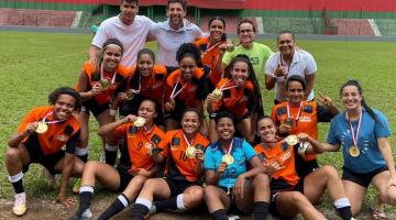 Jogadoras e comissão técnica mostram medalha com campo ao fundo #paratodosverem