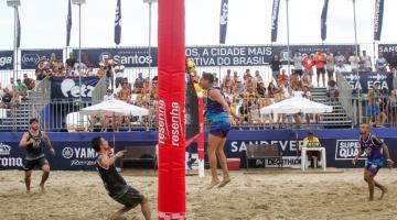 atletas estão jogando futvôlei na praia. Mastro da rede está ao centro da foto em primeiro plano. #paratodosverem