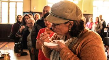 Mulher come um salgado com molho à base de café. Ela é a primeira de uma fila de pessoas. Com uma das mãos ela leva o salgado à boca e, com a outra ela segura um pequeno prato. #Pracegover