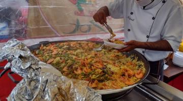 Cozinheiro, paramentado, retira alimentos de grande panela e coloca em prato. #paratodosverem