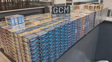 inúmeros pacotes com caixas de cigarro empilhados em sala. Placa com as letras gcm está sobre a pilha.  #paratodosverem