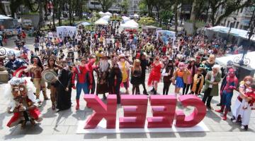 Cosplays ao lado de letreiro escrito geek e publico lotando a praça ao fundo #paratodosverem