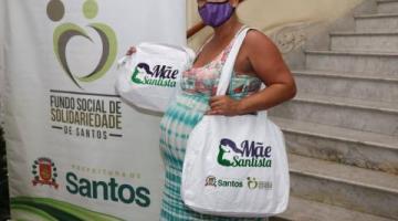 gestante está em pé ao lado de banner do fundo social de solidariedade. Ela segura sacola do programa mãe santista. #paratodosverem