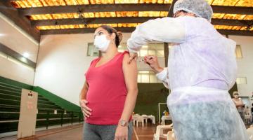GRAVIDA ESTÁ ACARICIANDO A BARRIGA ENQUANTO É VACINADA. A vacinação ocorre em um ginásio. #paratodosverem