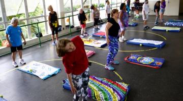 MULHERES ESTÃO FAZENDO EXERCÍCIO EM PÉ, AO LADO DE COLCHONETES. #PARATODOSVEREM