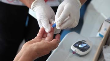 foto em close mostra mãos com luvas tirando gota de sangue da ponta do dedo de outra pessoa. Aparelho de medição de glicemia está ao lado. #paratodosverem